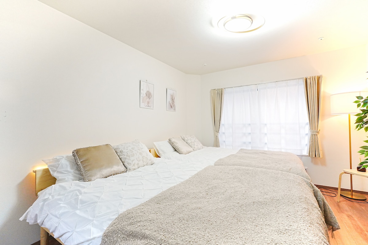 A serene sleeping area is presented, featuring two spacious double beds arranged side by side, with light-colored bedding and decorative pillows. Natural light filters through a sheer curtain, adding a soft glow to the room. Minimalist art decorates the walls, contributing to the calming ambiance.