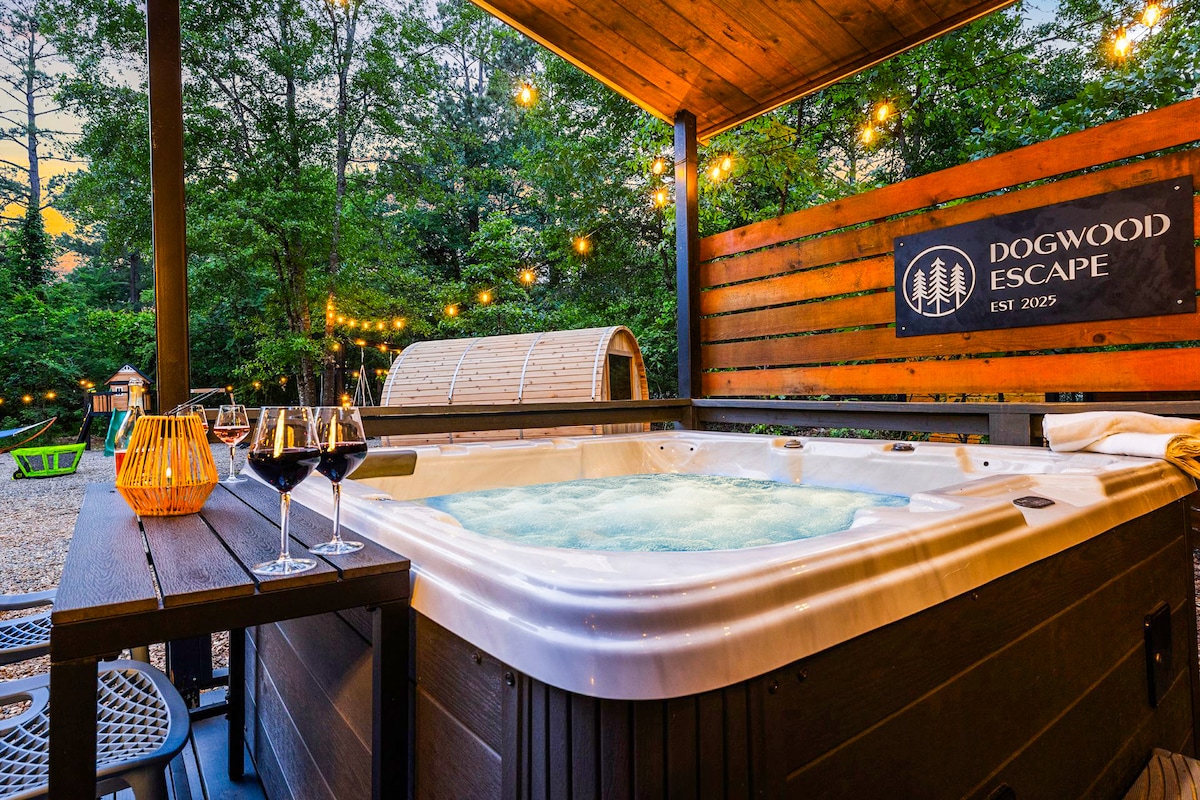 A hot tub is positioned on a wooden patio, surrounded by lush greenery and decorative string lights. Two wine glasses sit on a small table, hinting at a relaxing setting. The backdrop features wooden fencing and a sign reading 'Dogwood Escape'.