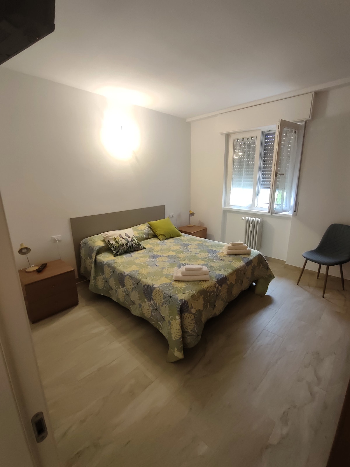 A well-lit bedroom features a spacious bed covered with a floral-patterned bedspread. Nightstands are located on either side of the bed, each with a lamp. A window allows natural light to illuminate the room, complemented by a simple chair in the corner.