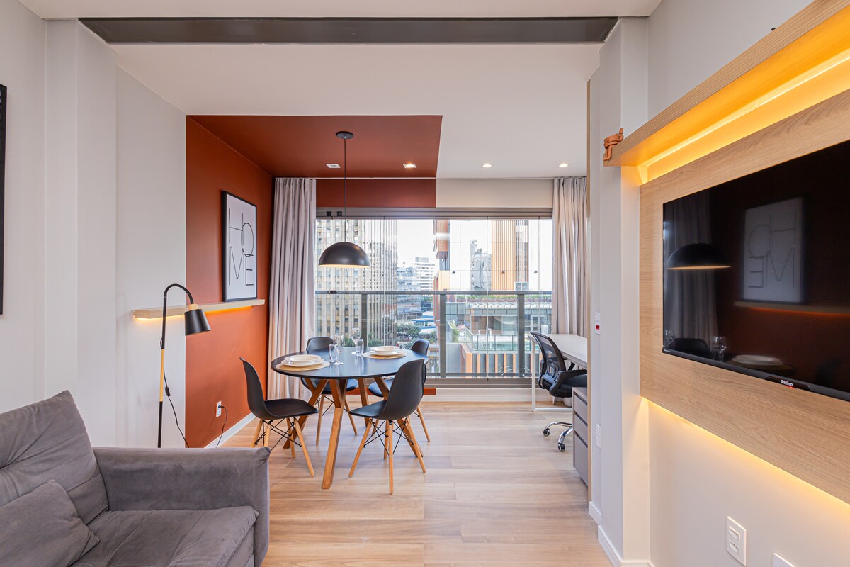 A modern living space features a comfortable sofa, a round dining table surrounded by chairs, and a workspace with a desk. Large glass doors provide access to a balcony, filling the area with natural light and offering views of the city skyline.