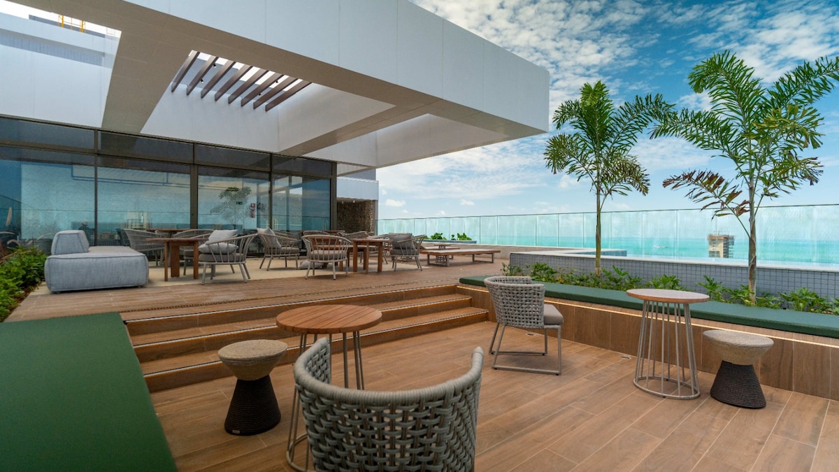 An outdoor rooftop area provides comfortable seating with modern furniture, including lounge chairs and tables. Lush greenery and palm trees frame the space, which offers a clear view of the horizon and the infinity pool reflecting the sky.