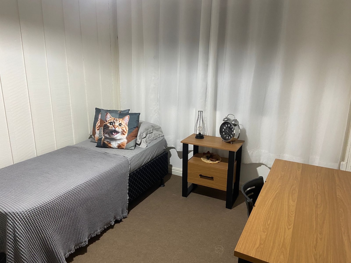 A simple bedroom is featured, showcasing a spacious bed topped with a textured gray blanket. A small desk and chair are positioned near a wooden dresser, alongside a decorative pillow with a cat image. Natural light filters through the sheer curtains.