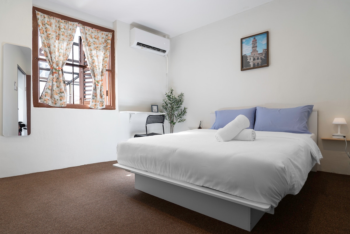 A spacious sleeping area features a comfortable bed with fresh linens. Natural light filters through floral-patterned curtains, illuminating the room. A small desk and chair are positioned near a large window, and an air conditioning unit is mounted on the wall.