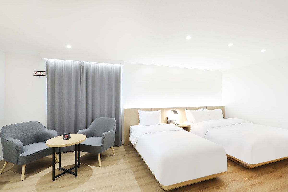 A well-lit room features two spacious beds with crisp white linens. Soft grey curtains frame a large window, allowing natural light to fill the space. A cozy sitting area is highlighted by a round table and comfortable chairs, providing an inviting spot for relaxation.