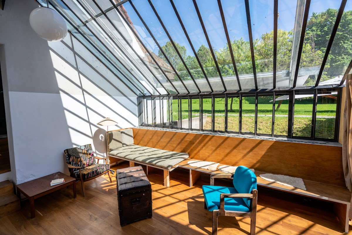 A bright, airy space is filled with natural light streaming through a large glass wall. A wooden bench runs along one side, complemented by colorful seating options. A small table is positioned nearby, creating an inviting area to relax and enjoy the surrounding greenery.