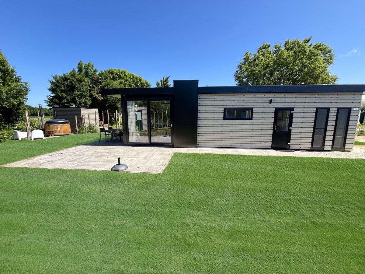 A modern chalet is set against a clear blue sky, featuring large glass doors that open to a patio. The spacious outdoor area is framed by well-maintained grass, with seating and a wood-fired hot tub visible in the background.