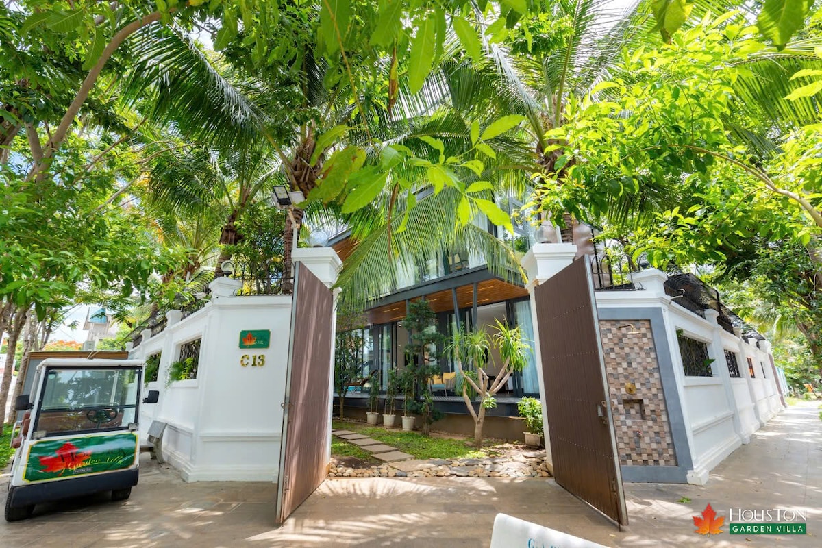 The entrance of the villa is framed by lush palm trees and vibrant greenery, creating a welcoming ambiance. A charming electric vehicle is parked nearby, and large glass windows reveal the contemporary design within. The gated entryway adds a sense of privacy.