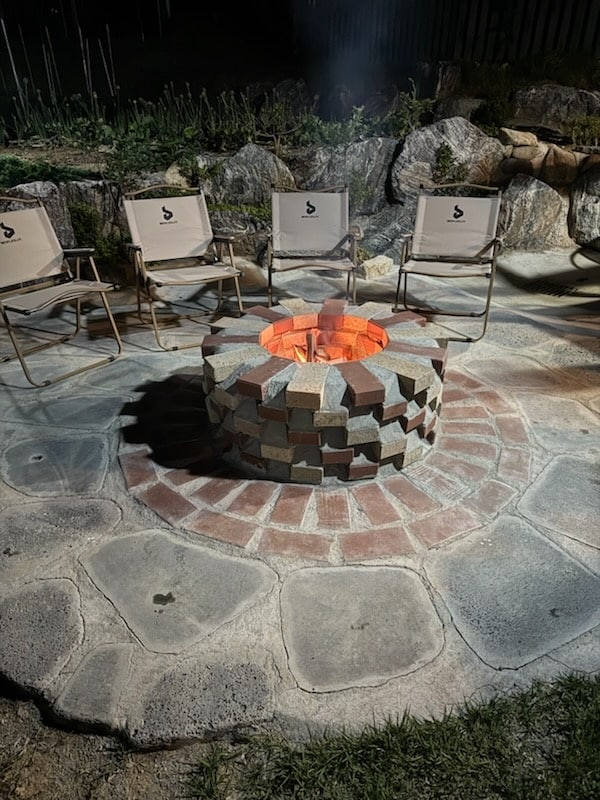 A circular fire pit is surrounded by several lightweight chairs made of sturdy fabric. A stone pathway and a mixture of natural and man-made materials create a rustic atmosphere. The bright glow of the fire is highlighted, casting warm light on nearby surfaces.