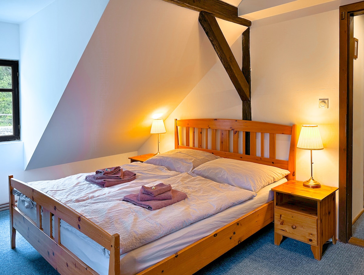 A bright and airy bedroom features a wooden bed with a spacious mattress, neatly arranged with folded towels. Soft lighting is provided by lamps on either side of the bed. Exposed wooden beams complement the angled ceiling, creating a cozy atmosphere.