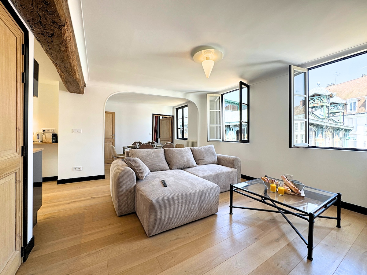 The living area features a spacious corner sofa in soft fabric, complemented by a glass coffee table. Large windows provide ample natural light and reveal views of the surrounding architecture. Wooden beams add character to the room, enhancing its inviting atmosphere.