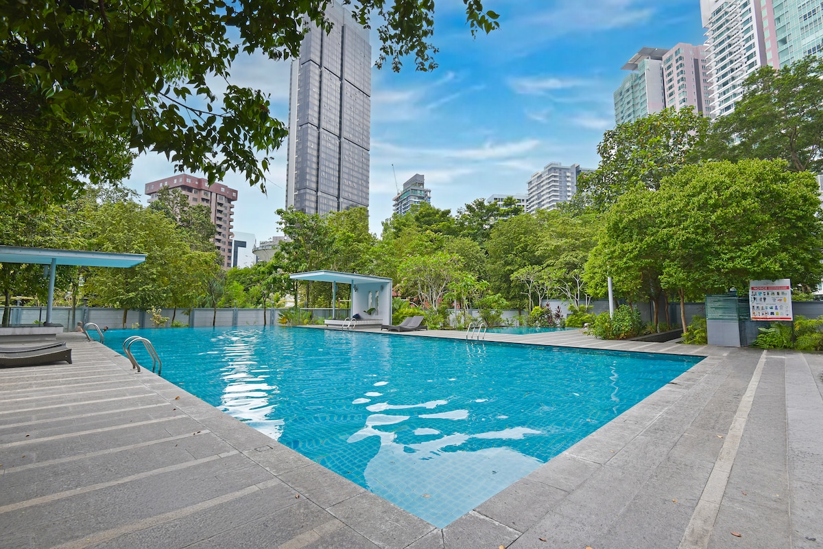 An outdoor swimming pool is surrounded by lush greenery and modern buildings. The pool features clear blue water and accessible steps for entry. Lounge areas with shaded seating are seen along the perimeter, creating a serene spot within the urban landscape.
