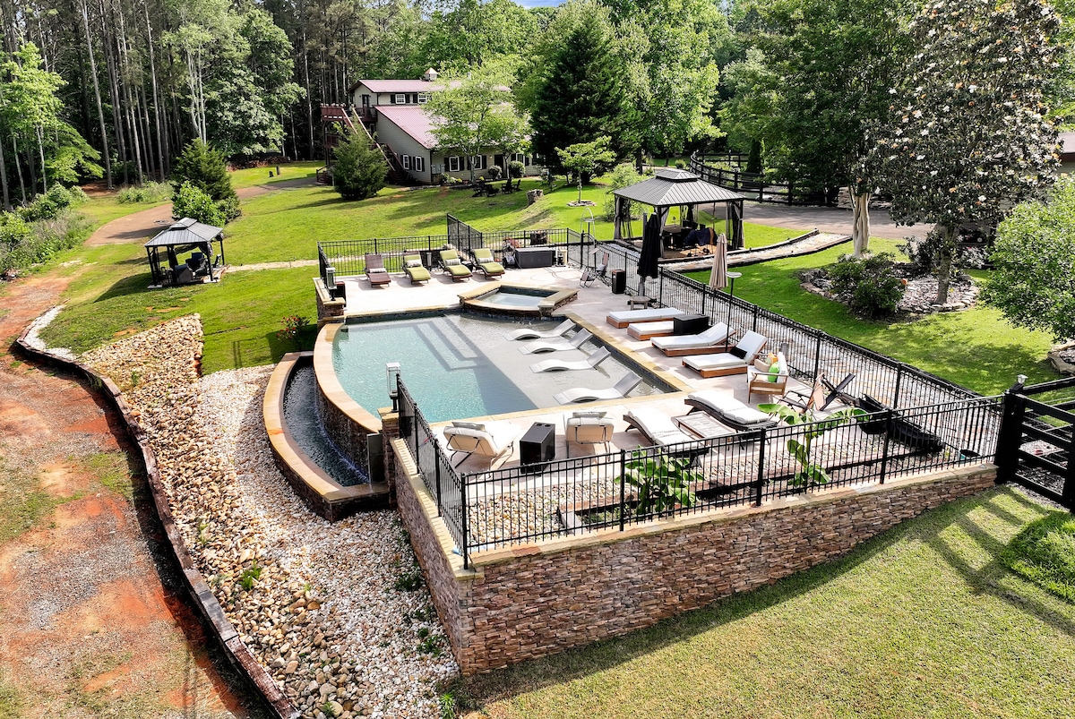 An aerial view captures the expansive outdoor area, featuring a private pool surrounded by lounge chairs and a stone landscape. Lush greenery and trees frame the setting, with additional seating and shaded structures visible in the distance.