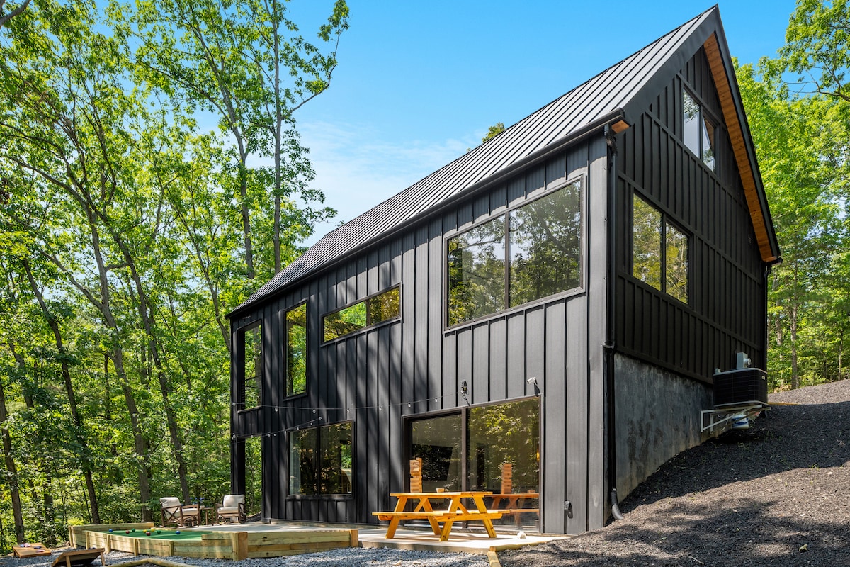 A modern cabin sits nestled among lush trees, featuring a striking black exterior. Large windows allow natural light to fill the interior, while a spacious deck with a picnic table provides an inviting outdoor gathering space for guests.