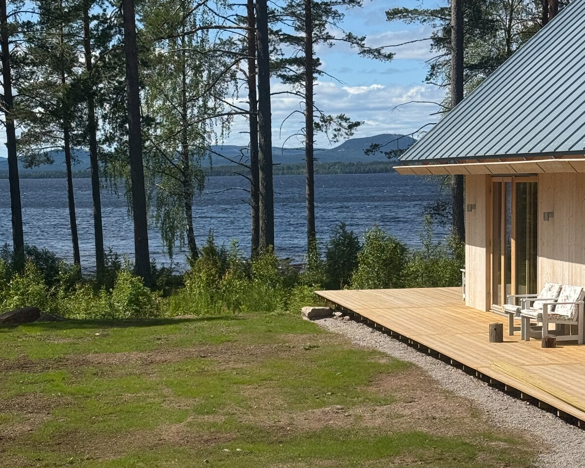 A serene view of the lake is captured, framed by tall trees and distant mountains. The wraparound deck features minimalist seating, inviting relaxation. Sunlight reflects off the water, while a lush, green lawn leads to the shoreline, creating a peaceful outdoor environment.