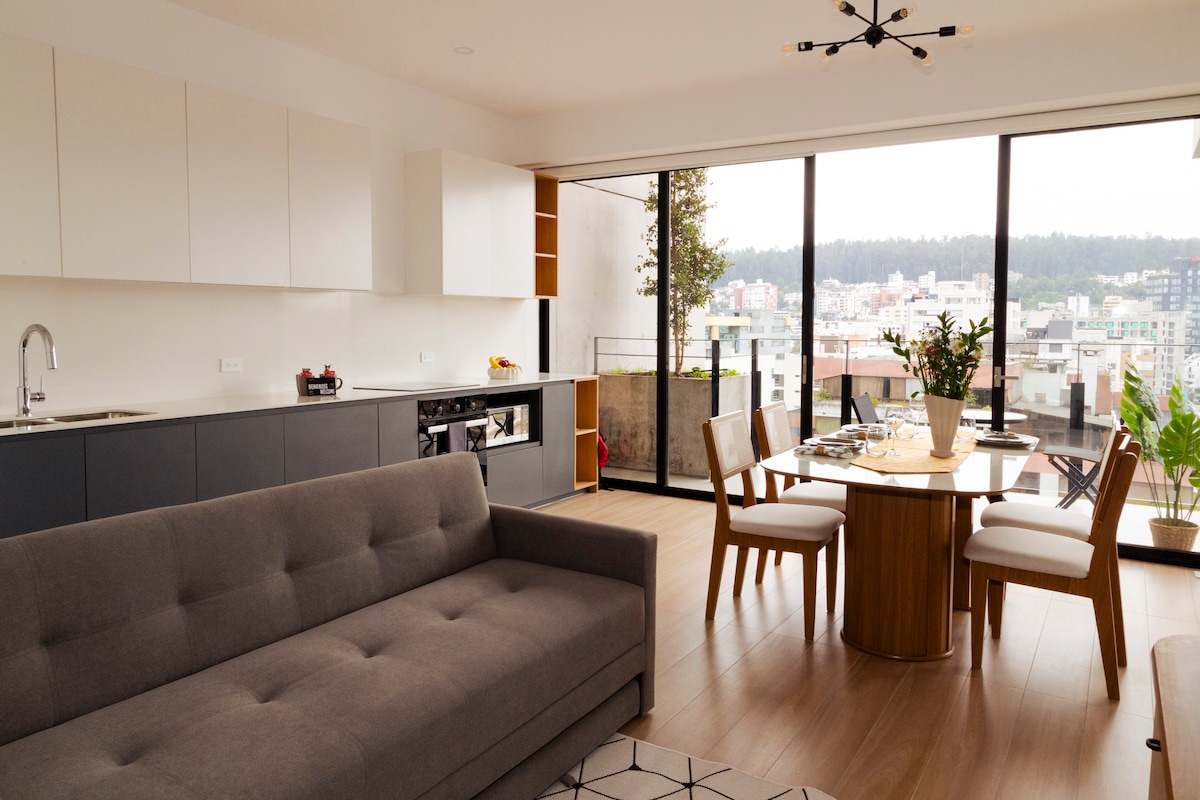 A modern living area features a spacious gray sofa positioned next to a large window. A dining table with four chairs is displayed, adorned with a decorative centerpiece. Natural light floods the space, highlighting the sleek cabinetry and open layout.