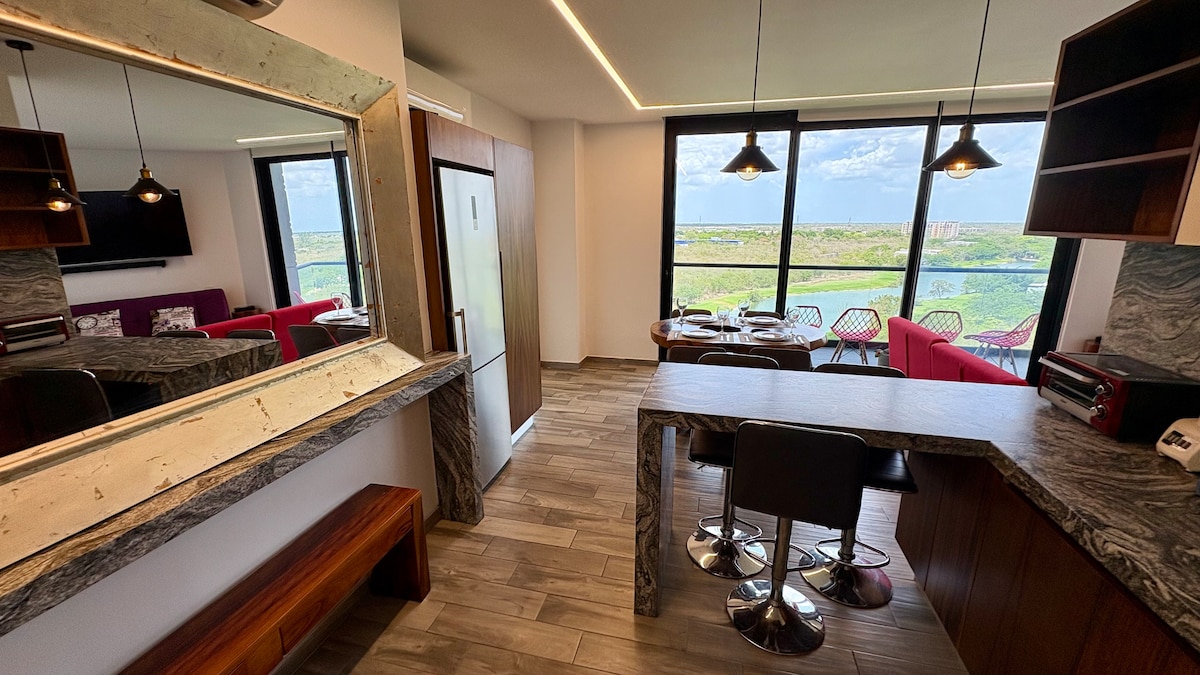 A modern kitchen and dining area are featured, with sleek marble countertops and stylish barstools. Large windows offer a scenic view of the surrounding landscape, while a dining table is positioned nearby. A cozy living area with a flat-screen TV is also visible.