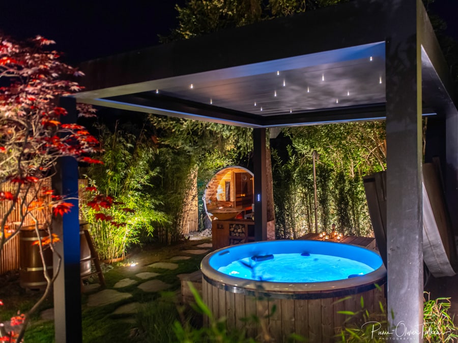 A jacuzzi is situated beneath a star-lit pergola, surrounded by lush greenery and softly glowing lights. Pathways of stones lead to a wooden sauna, creating a serene and inviting atmosphere for relaxation.