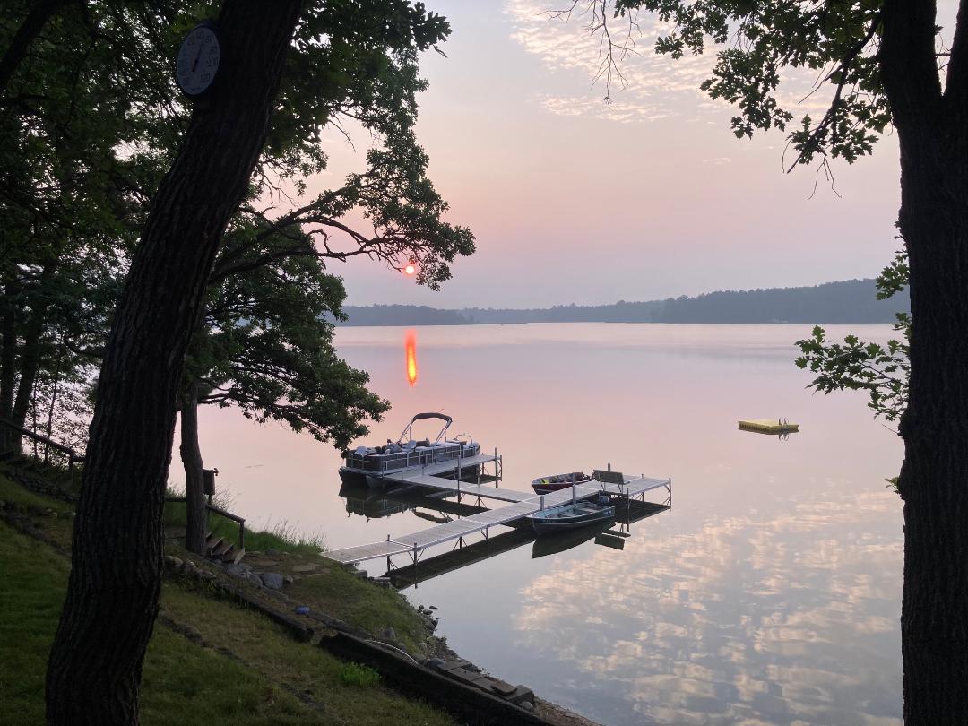 A serene lake view features calm waters reflecting the sunset, framed by trees on both sides. A dock extends into the lake, with two boats moored nearby. The horizon displays soft hues from the setting sun, creating a peaceful atmosphere.