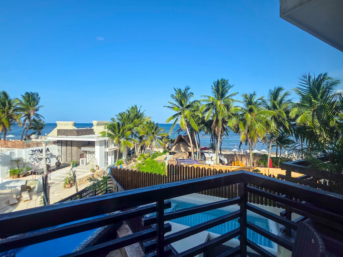 A stunning ocean view is captured from a balcony, overlooking palm trees and the vibrant beach area. The turquoise waters can be seen in the background, complemented by bright blue skies, creating a serene coastal atmosphere.