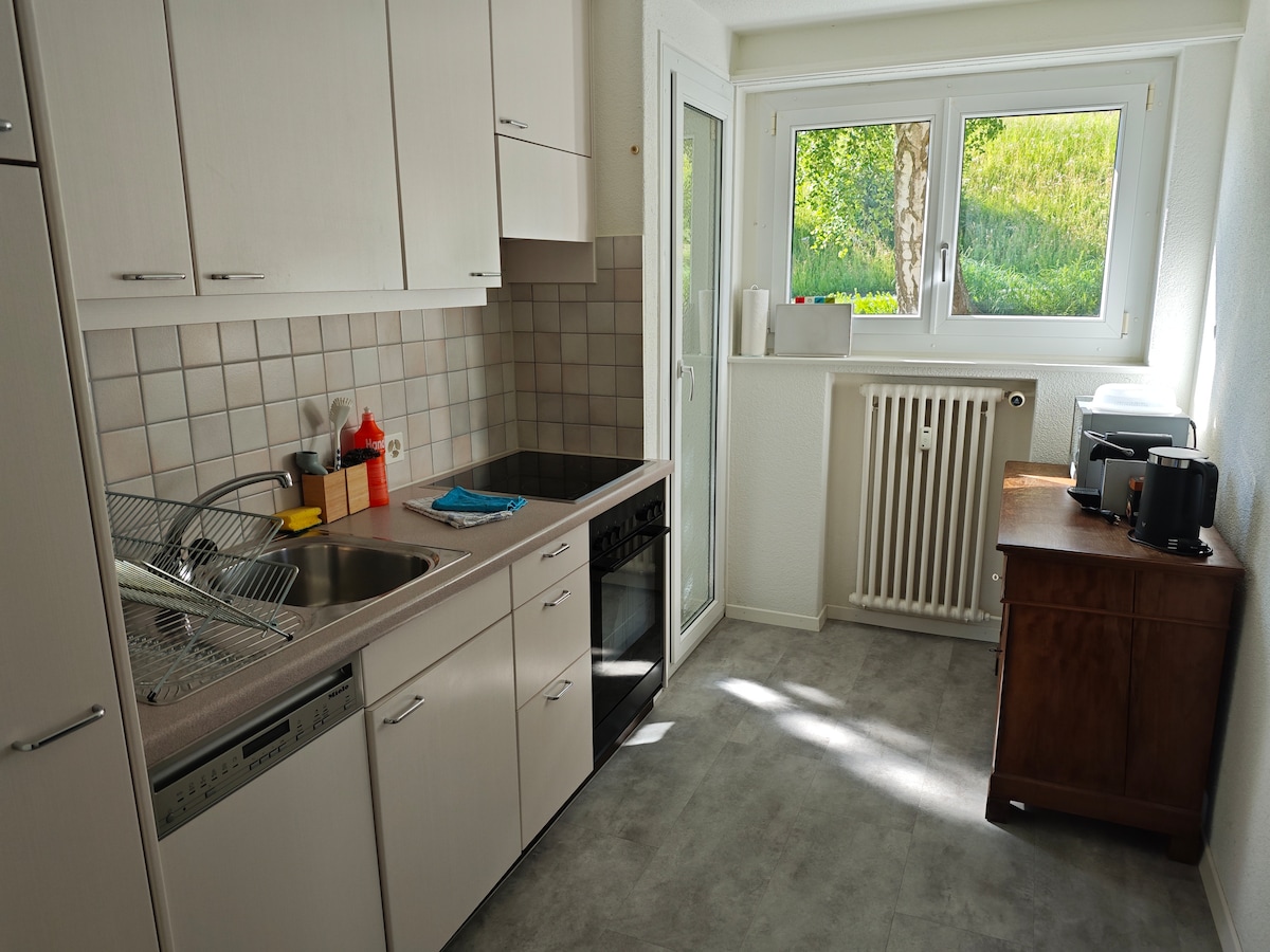 The kitchen space is equipped with essential appliances, including an oven, stovetop, and dishwasher. Counter space is complemented by cabinets and a sink. Natural light flows in through large windows, while a small wooden cabinet is placed against the wall.