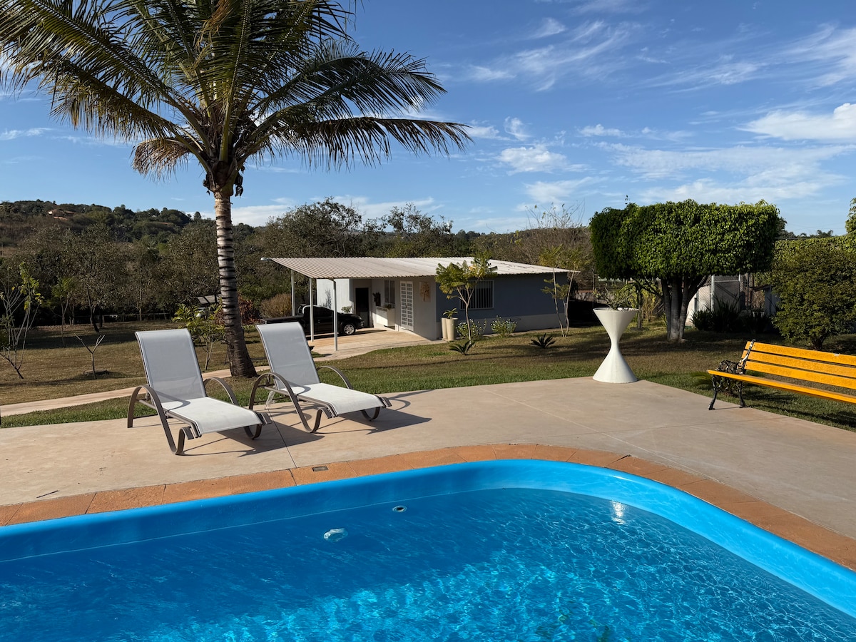 A private pool with clear blue water is framed by two lounge chairs on a sunlit patio. In the background, a house with a shaded porch is surrounded by green grass and trees under a clear sky.