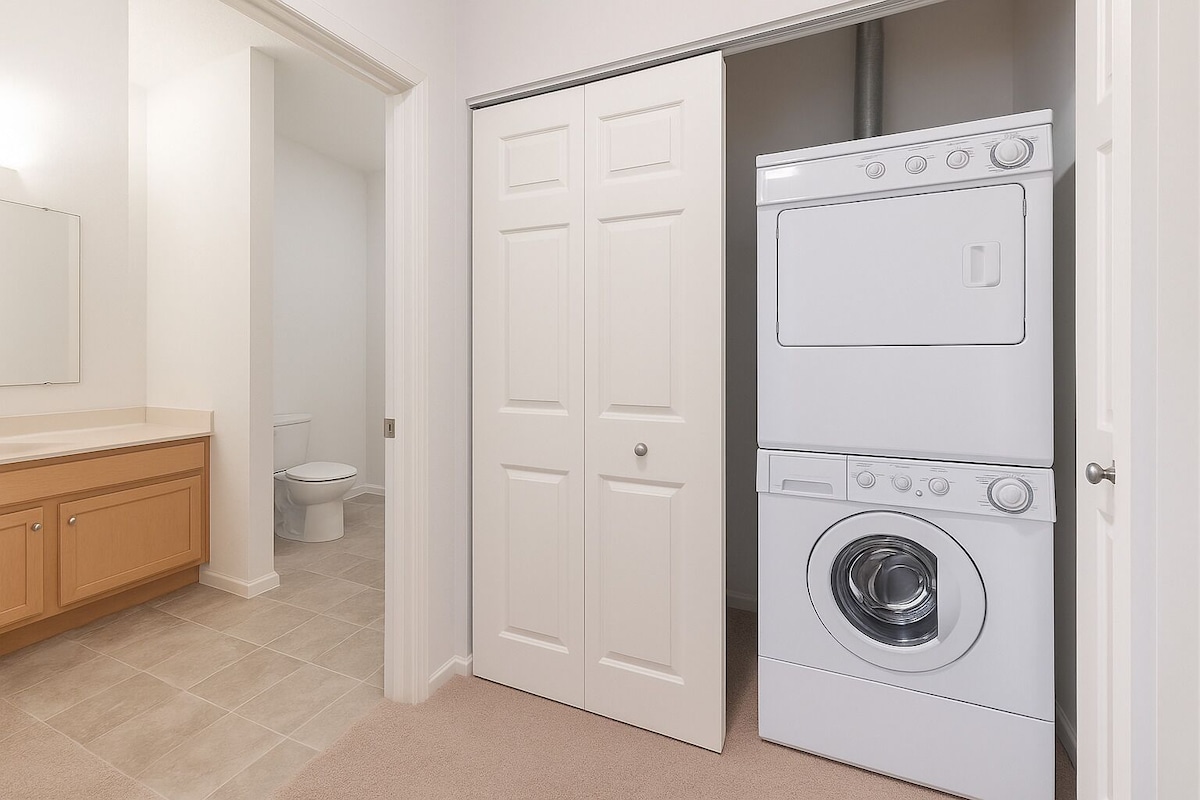 A laundry area is presented within the bathroom, featuring a stacked washer and dryer unit. Light tan cabinetry and a mirror are visible, while a toilet is discreetly positioned in the background. The flooring consists of large tiles, contributing to a clean and functional space.