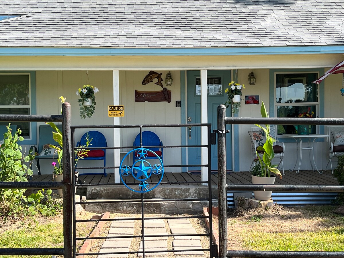 A welcoming entrance features a covered front porch with two blue chairs and a white table. Hanging plants and decorations add charm. A wrought iron gate leads to the pathway, which has stepping stones bordered by greenery and a small decorative sign.