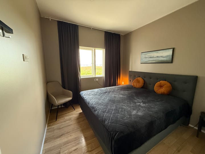 Este acogedor dormitorio cuenta con una cama de 180 cm de ancho con un cabecero gris acolchado y una funda acolchada a juego. Las paredes de color topo claro complementan el suelo de madera y las cortinas grises, mientras que los muebles minimalistas y un cuadro de paisaje sereno aportan calma y estilo.