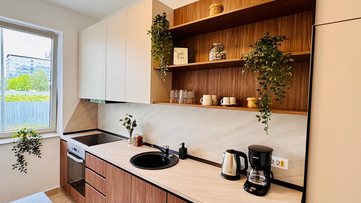 A modern kitchen features a variety of cabinets and an open shelf with plants and decorative items. A black circular sink and a coffee maker are visible, along with a stove and an oven. Natural light enters through a window, enhancing the warm oak tones of the cabinetry.
