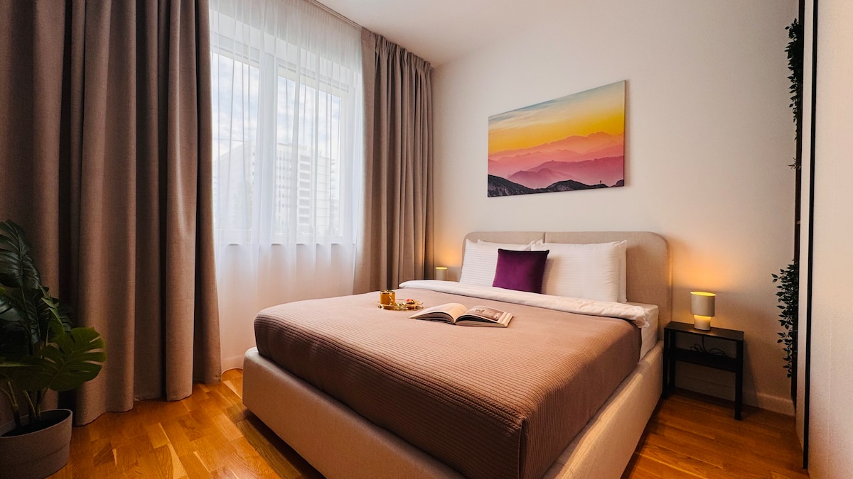 A spacious bedroom features a large bed with soft linens, complemented by bedside tables with lamps. Natural light filters through sheer curtains, illuminating a wall adorned with a landscape painting. A small plant adds a touch of greenery to the inviting space.