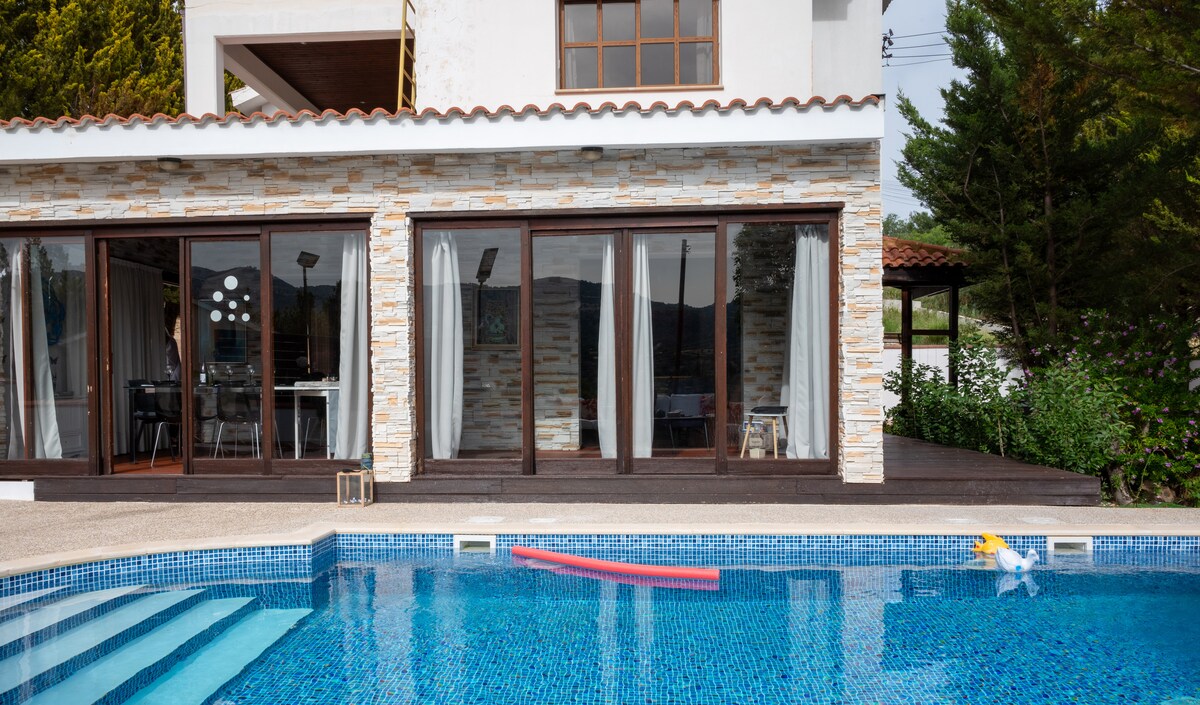 A private pool is framed by a spacious wooden deck, offering an inviting atmosphere. Large glass sliding doors reveal views of the modern interior. The exterior is complemented by stone accents and lush greenery, providing a serene setting for relaxation.