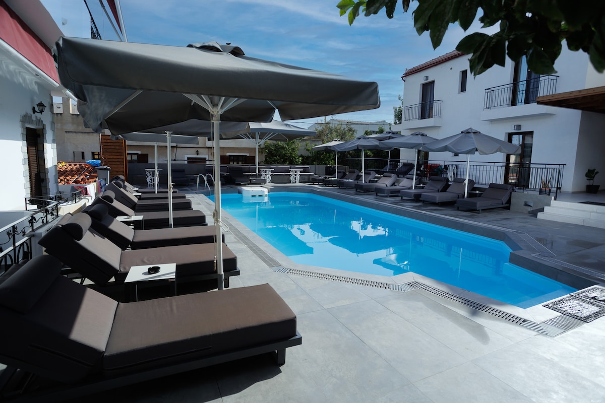 A tranquil pool area features a rectangular swimming pool surrounded by comfortable loungers. Grey umbrellas provide shade, while potted greenery adds a touch of nature. A spacious terrace is visible in the background, contributing to an inviting outdoor setting.