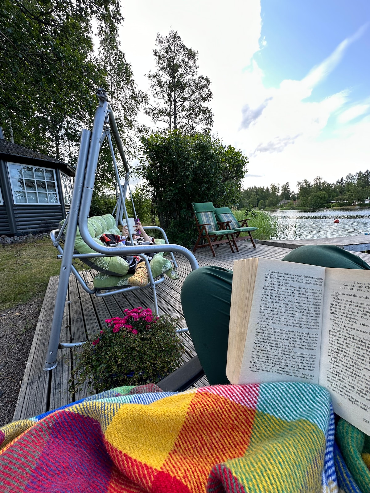 A seating area by the water is depicted, featuring comfortable chairs and a swing with cushions. A colorful blanket covers part of the view, while a book is being read. Lush greenery and a peaceful lake can be seen in the background.