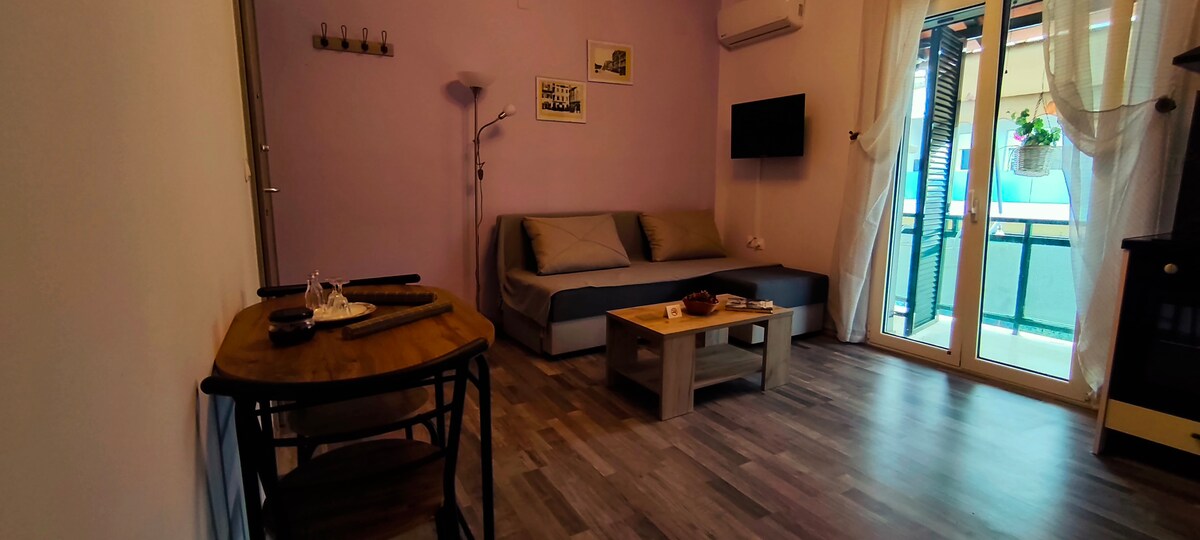 A cozy living area features a plush sofa positioned against a light purple wall. A smart TV is mounted on the opposite wall, while natural light flows through large glass doors leading to a balcony. A wooden dining table offers space for two, accompanied by decorative items.