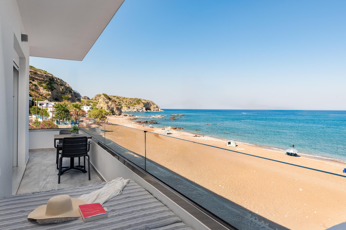 A spacious balcony offers stunning views of the beach and sea. Sunbeds are arranged for relaxation, with a hat and book casually placed on a soft blanket. The shoreline features soft sand with boats gently bobbing in the clear water under a bright blue sky.