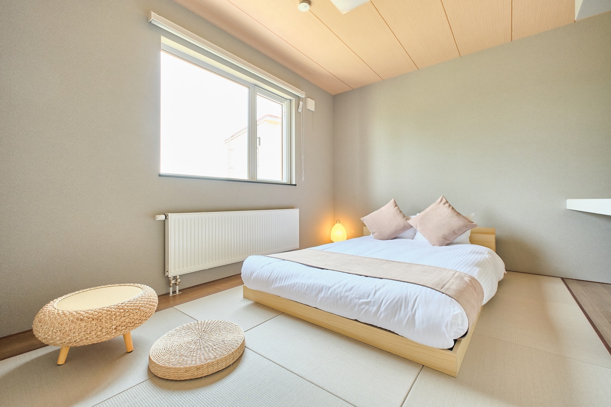 和洋室／ベッドルーム。畳の心地よさを感じる空間に、ダブルサイズのローベッドを配置。清潔な寝具と木目調の天井が、格別なリラックスタイムを提供します。大きな窓とムードのあるランプで、ゆったりお過ごしください。