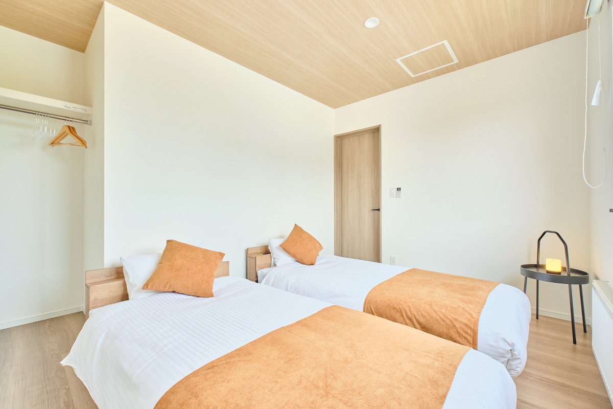 ベッドルーム。木目調の天井が温もりを感じさせる広々とした空間です。シングルベッド2台と、衣類を掛けられるオープンクローゼットを完備。清潔な寝具と落ち着いたトーンの内装が、心からリラックスできる快適な滞在を提供します。