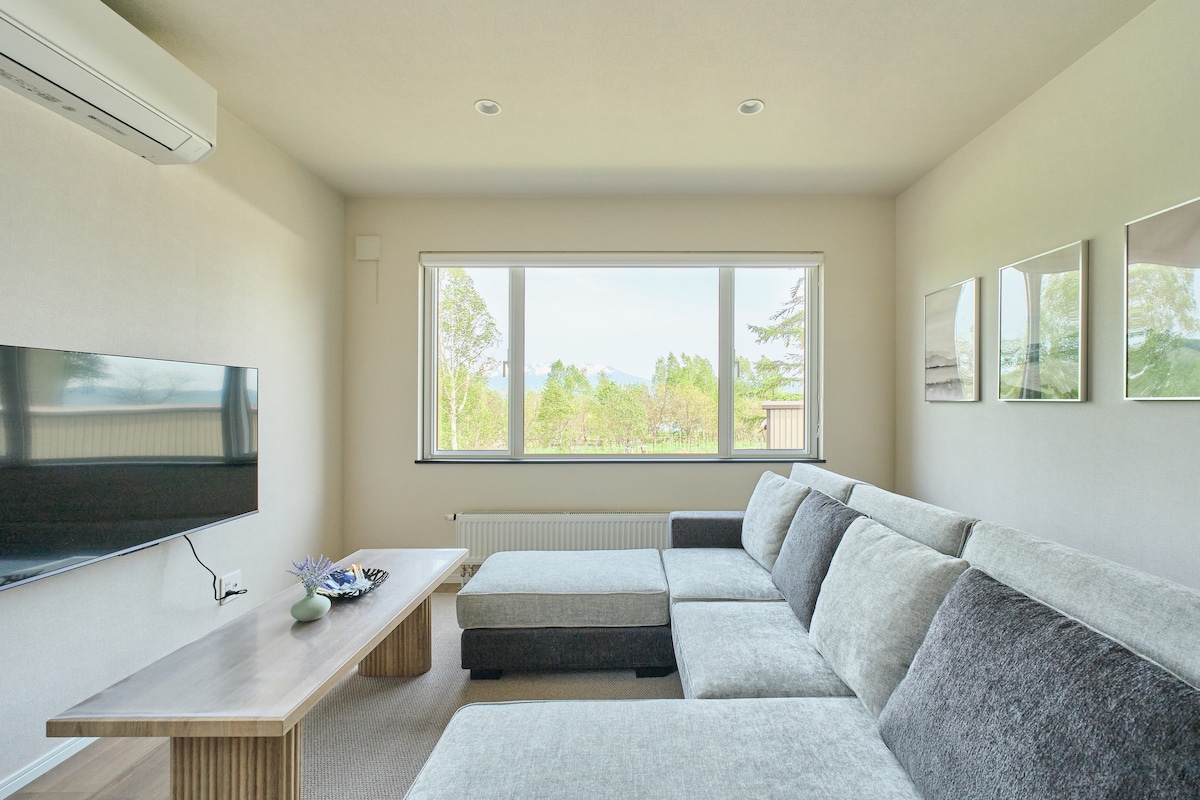 自然光が差し込む広々としたリビングルームです。窓からは豊かな緑の景色が広がり、リラックスできます。足を伸ばして寛げるL字型大型ソファと大画面TVを完備。清潔感溢れる空間で、グループやご家族の滞在を快適にサポートします。