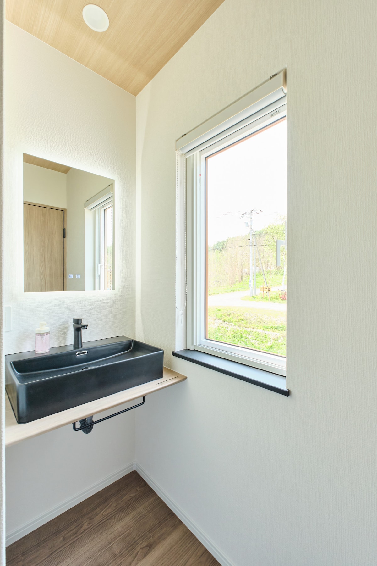 木目と白壁が調和したナチュラルな空間。窓から自然光が差し込み、明るく清潔感があります。目を引く黒いスクエア洗面ボウルとLEDバックライト付きミラーで、朝の身支度もスタイリッシュに。