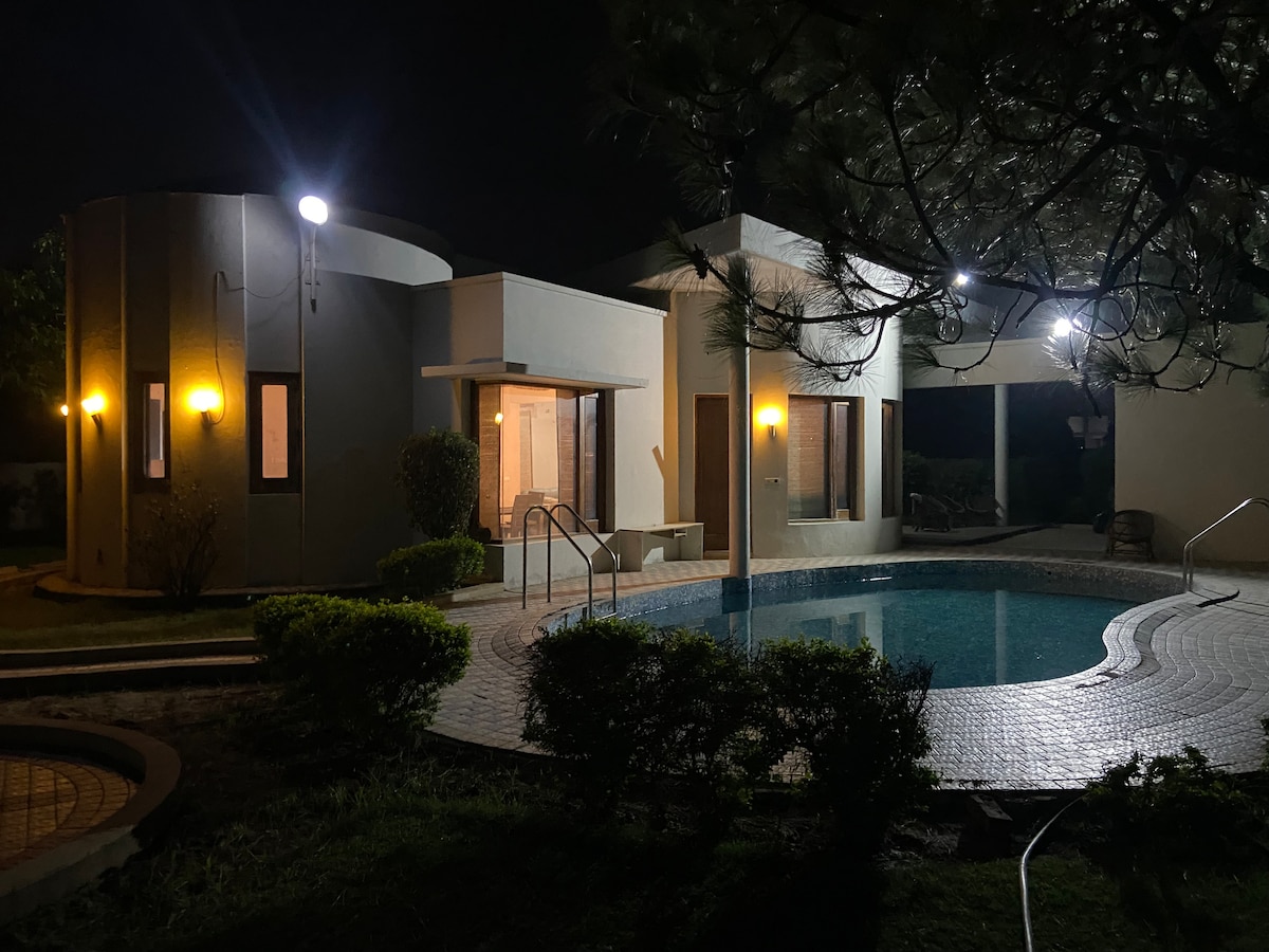 A modern accommodation is set against a nighttime backdrop, featuring soft yellow lights illuminating the exterior. A circular pool, surrounded by landscaping, reflects the surrounding structure, creating a serene ambiance. The entrance showcases ample glass windows, enhancing visibility into the living spaces.