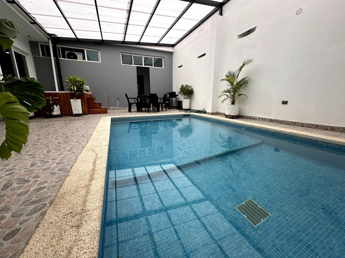 A modern swimming pool reflects the surrounding light, situated within a minimalist courtyard. Potted plants are positioned around the pool's edge, and a dining area with a table and chairs is visible in the background, along with a small set of steps leading to an elevated space.