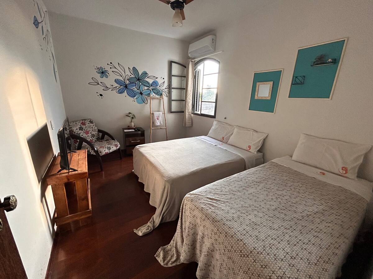 The guest room features two spacious beds covered with light-colored bedspreads. A comfortable chair is placed in the corner. Wall art with blue flowers adds a decorative touch, while natural light filters through a window adorned with shutters.