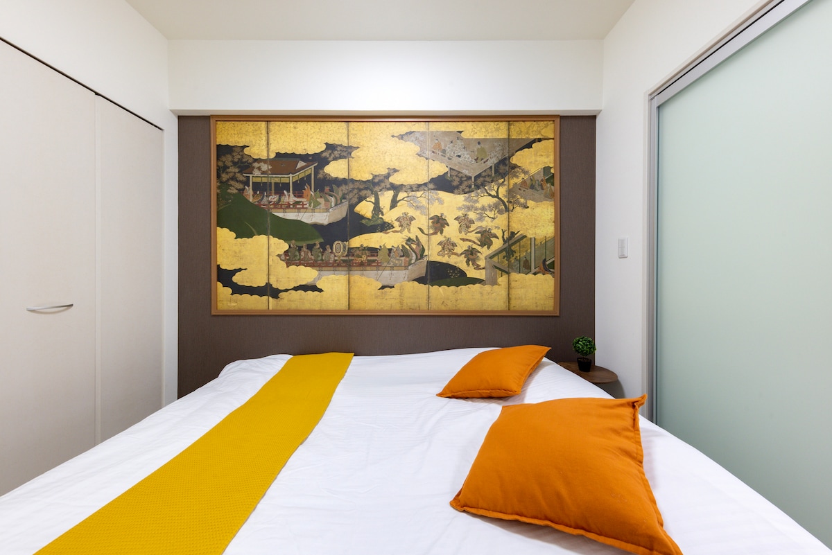A comfortable sleeping area features a spacious bed adorned with white linens and two orange accent pillows. A large, intricate wall art piece depicting a traditional landscape scene serves as a striking focal point above the bed, enhancing the room's overall ambiance.