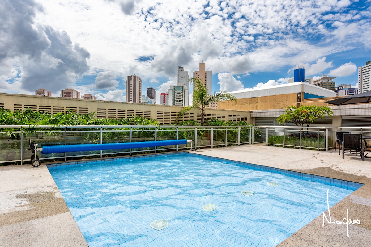 The image displays a rooftop swimming pool with clear blue water under a partly cloudy sky. Lush greenery surrounds the pool area, and a lounge chair is positioned nearby. A long blue bench runs along one side, enhancing the serene setting.