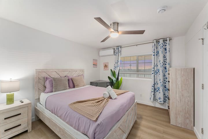 Оваа светла спална соба има рустикален дрвен кревет со мека прекривка за кревет од лаванда и соодветен мебел. Огледален плакар, додека сиво биро создава работен простор. Големите прозорци, таванскиот вентилатор и клима уредот обезбедуваат удобност и природна светлина.
