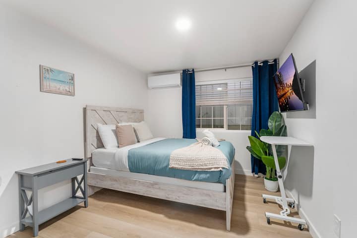 Оваа удобна спална соба има рустикална дрвена рамка за кревет со син јорган и беж плетенка. Ноќно шкафче што одговара со ламба и голем прозорец. Ѕиден телевизор, саксиско растение и минимален декор создаваат модерен, привлечен простор