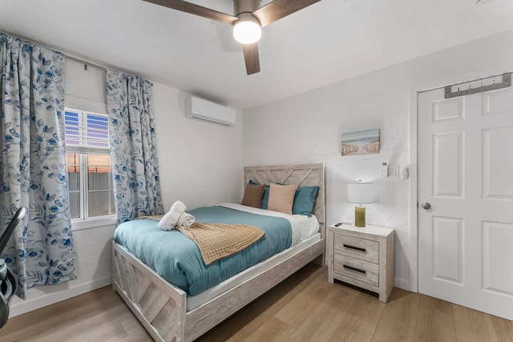 Оваа модерна спална соба инспирирана од крајбрежјето има рустикален дрвен кревет со сина прекривка за кревет, беж фрлање и соодветна ноќна масичка. Огледалниот плакар и стол создаваат работен простор. Тропската уметност, растението во саксија и вентилаторот на таванот додаваат стил и удобност.