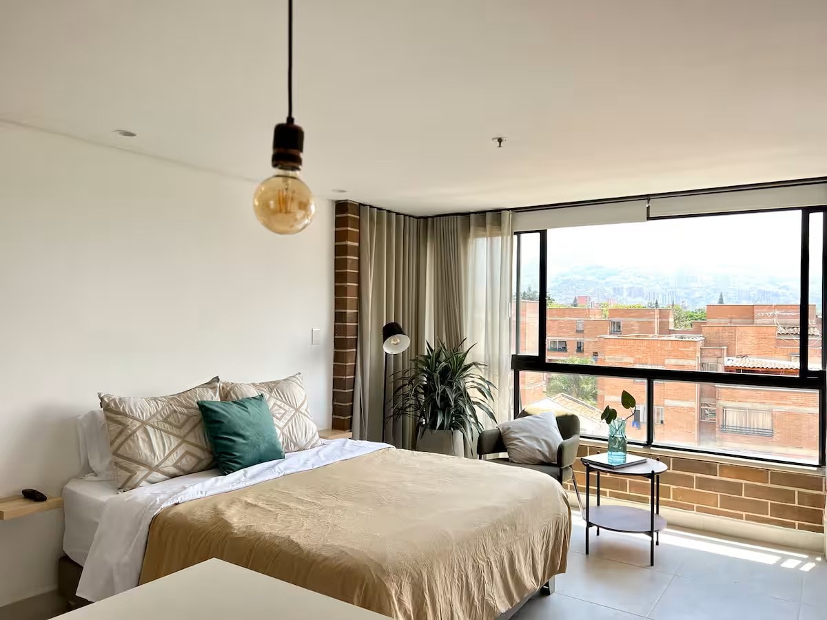 A spacious bedroom features large windows offering views of the surrounding area. The bed is dressed in neutral-toned linens, complemented by a decorative pillow. A reading lamp and a small side table are situated next to a cozy armchair, accompanied by a potted plant.