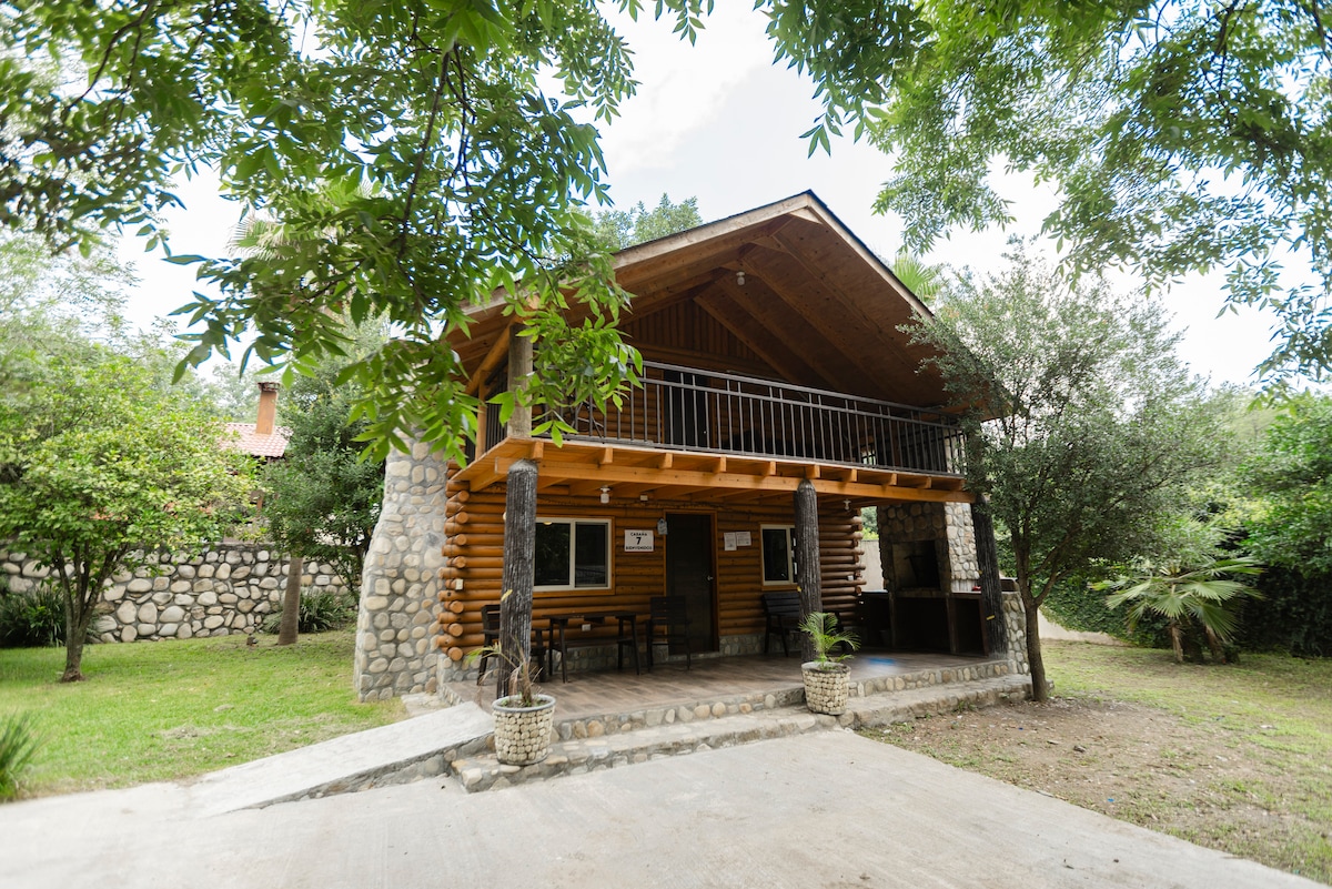A wooden cabin is situated beneath a canopy of trees, featuring a spacious covered porch. The exterior showcases stone accents and a warm wood finish, surrounded by a well-maintained lawn and natural landscaping. The upper balcony overlooks the outdoor area.