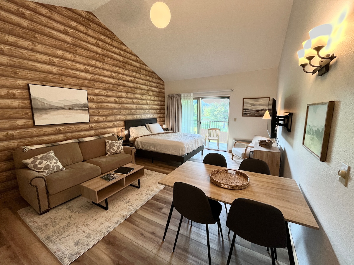 A studio condo is presented with a warm wood-paneled accent wall. A spacious bed is positioned alongside a sofa, with a dining table set for four. Large windows illuminate the room, providing views of the balcony and surrounding nature.